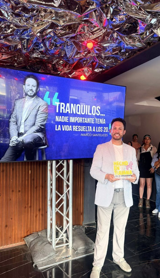 El empresario Marco Antonio Moreno Gómez Santelices posa sonriente sosteniendo su reconocimiento tras su participación en la Feria de Emprendimientos Hecho en Tijuana frente a una pantalla con su fotografía y frase motivacional.