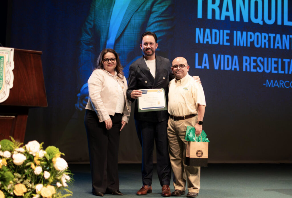 Marco Santelices sosteniendo un reconocimiento de la FCA UABC junto a autoridades universitarias en el escenario tras su conferencia por el 65 aniversario.